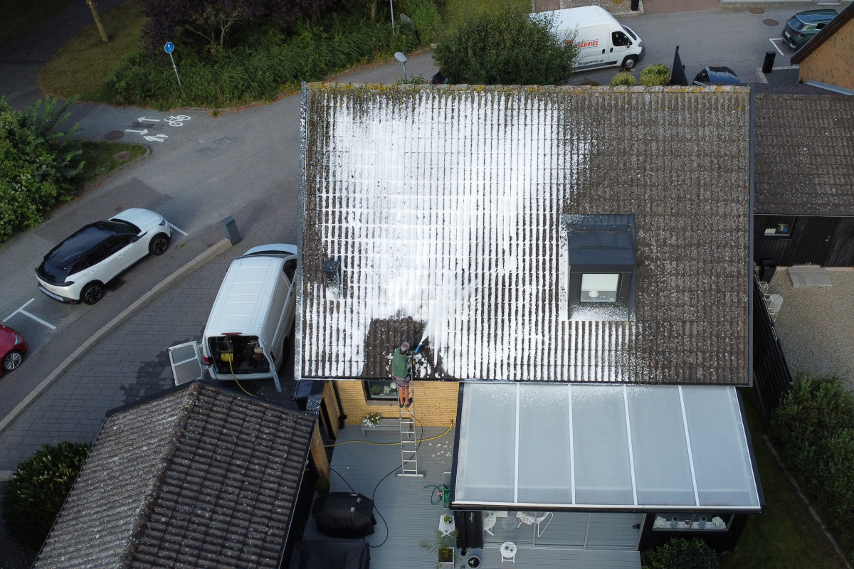 En takbehandlare arbetar på ett hustak som är delvis täckt med ett skummande rengöringsmedel. En vit skåpbil står parkerad bredvid huset, och en stege är lutad mot väggen. Det omgivande området inkluderar bilar och en gångbana med cykelmarkeringar.